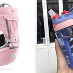 Pink baby carrier and blue water bottle with red lid held by a hand against a blurred background.