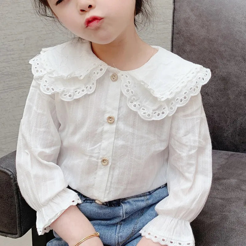 Child wearing a white blouse with lace details and blue jeans sitting on a gray couch.