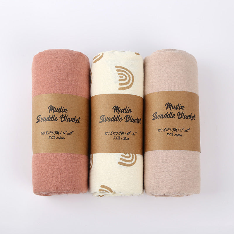 Three muslin swaddle blankets in different colors with brown labels on a white background