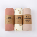 Three muslin swaddle blankets in different colors with brown labels on a white background