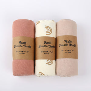 Three muslin swaddle blankets in different colors with brown labels on a white background