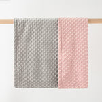 Two textured baby blankets, one gray and one pink, on a wooden rod against a white background.
