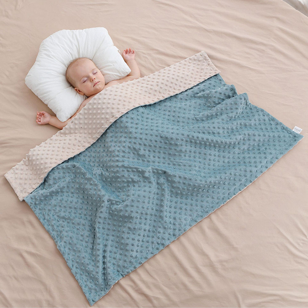 Baby sleeping on a beige blanket with a blue textured blanket draped over it, on a beige bed.