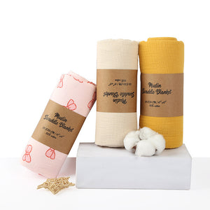 Three muslin swaddle blankets in pink, beige, and yellow with brown labels on a white background.