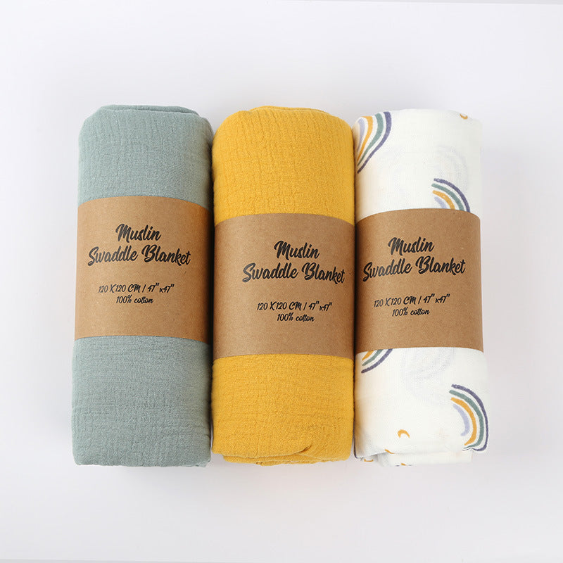 Three swaddle blankets in green, yellow, and white with brown labels on a white background