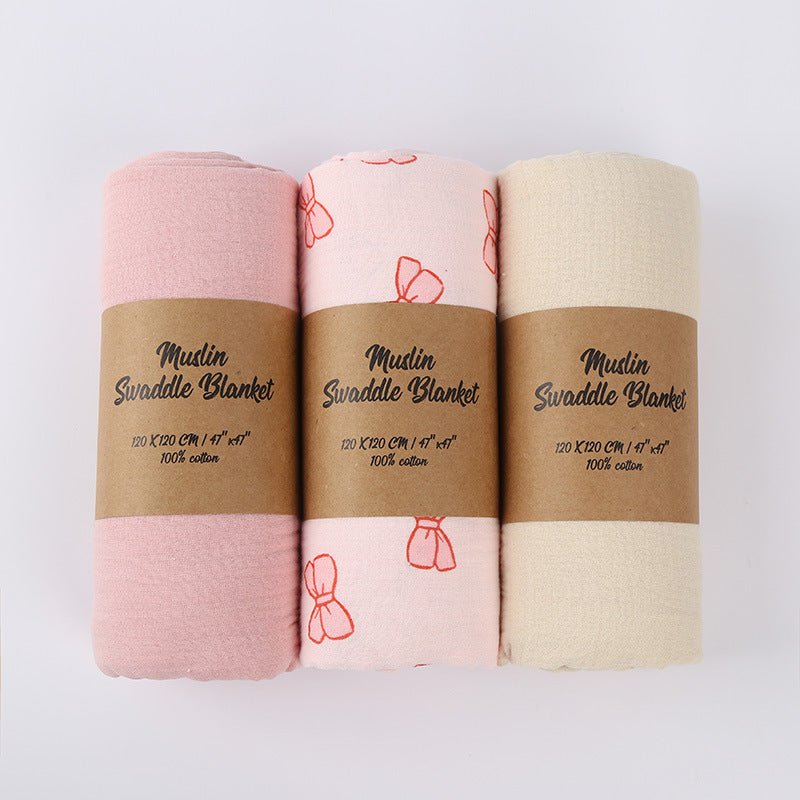 Three muslin swaddle blankets in pink, white, and beige with brown labels on a light gray background.