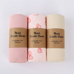 Three muslin swaddle blankets in pink, white, and beige with brown labels on a light gray background.