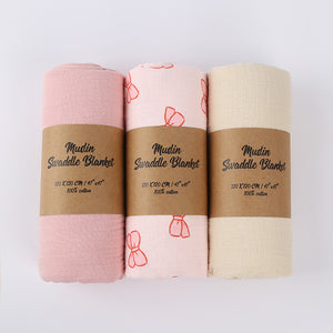 Three muslin swaddle blankets in pink, white, and beige with brown labels on a light gray background.
