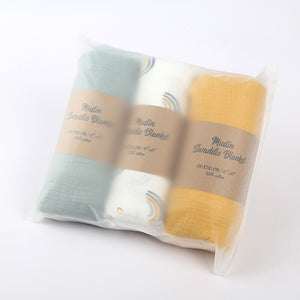 Set of three muslin swaddle blankets in green, white, and yellow packaging on a white background