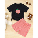Black t-shirt with a red apple design and red checkered shorts on a wooden surface.