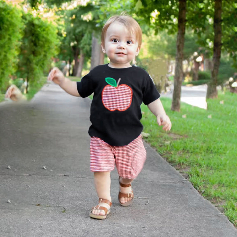 Child wearing a black shirt with a red apple graphic and pink shorts walking on a sidewalk.