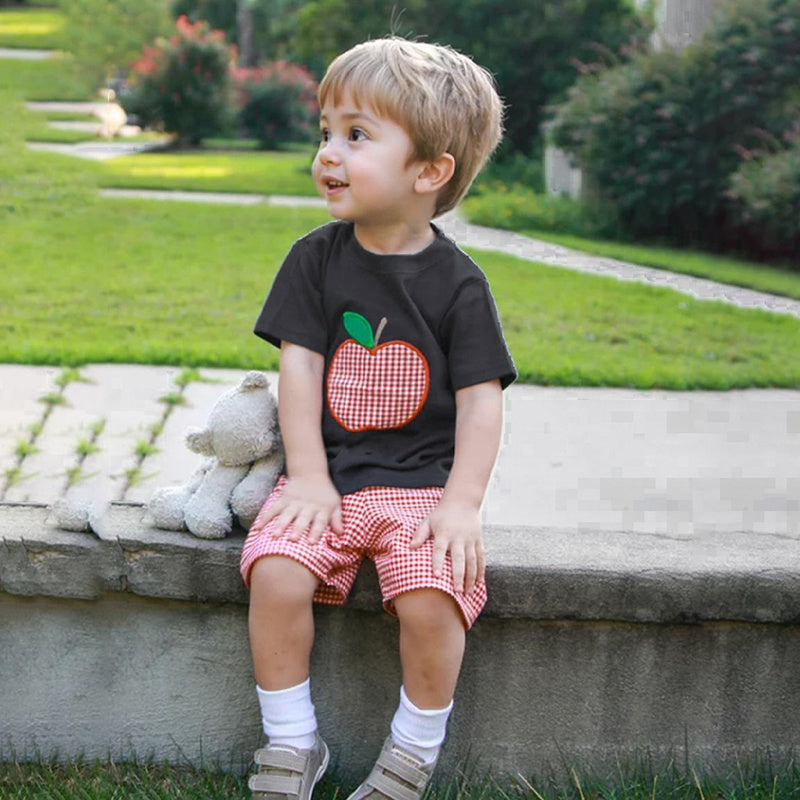 Child wearing a black t-shirt with a red apple graphic and red checkered shorts, sitting on a stone ledge outdoors.