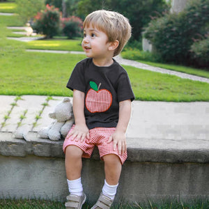 Child wearing a black t-shirt with a red apple graphic and red checkered shorts, sitting on a stone ledge outdoors.