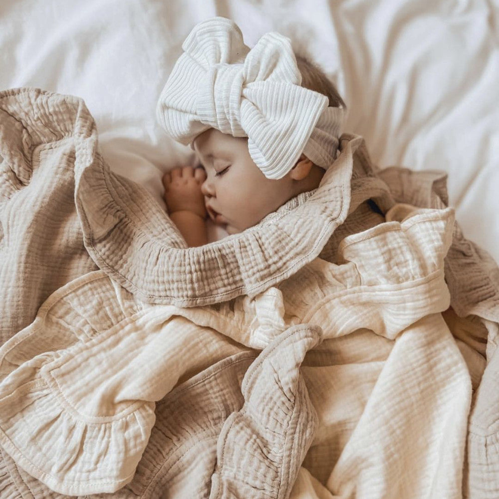 Baby wrapped in a beige blanket with a large white bow headband, lying on a soft surface.