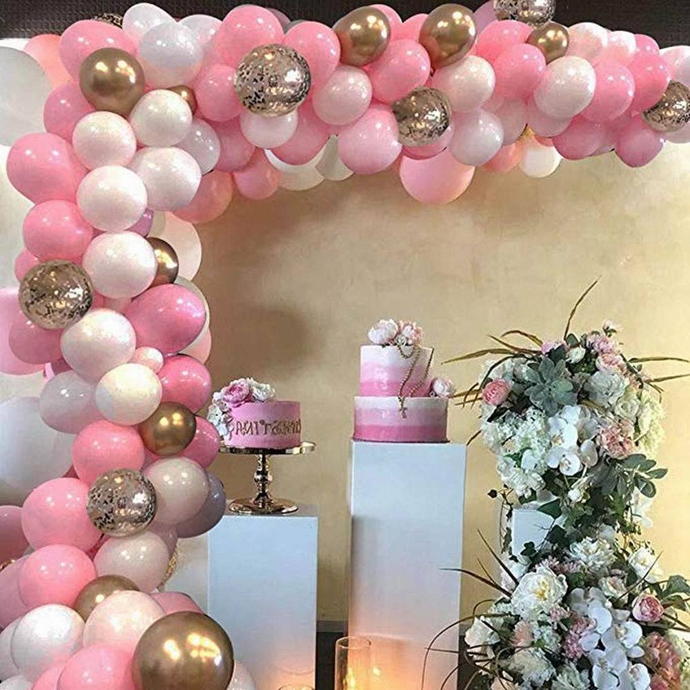 Balloon arch with pink, white, and gold balloons, two cakes on stands, and floral decorations against a beige wall.