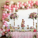 Decorative balloon arch with pink, white, and gold balloons, flowers, and a cake on a table.