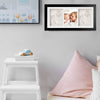 Framed baby handprints and footprints on a wall above a white shelf with a toy car and pink cushions.