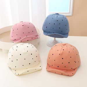 Four polka dot baby caps in pink, blue, white, and peach on a light surface.