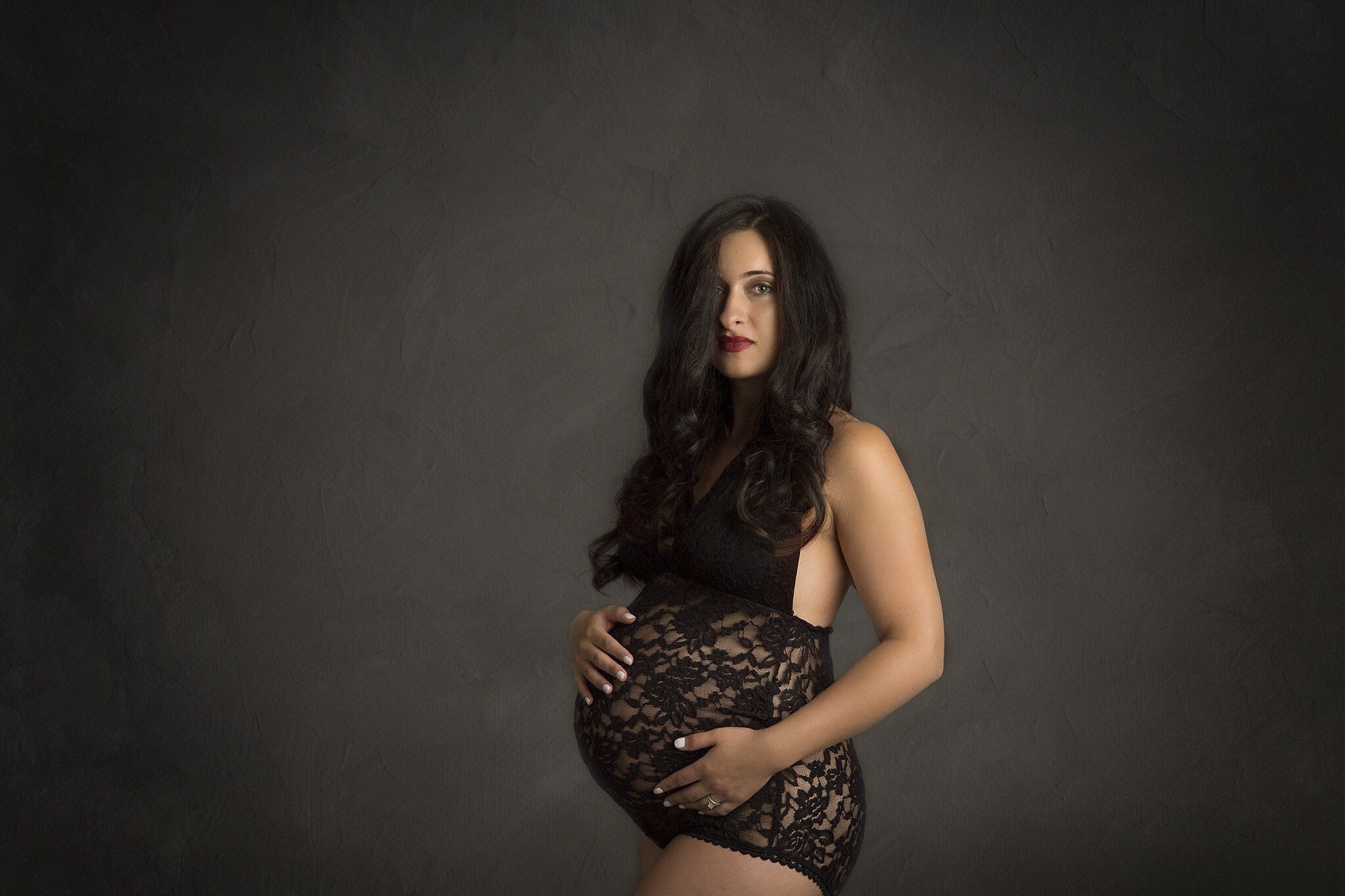 Pregnant woman wearing a black lace bodysuit against a dark background