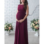 Woman wearing a burgundy maternity dress with floral lace top against a white brick wall.