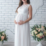 Woman in a white maternity dress standing in front of a white brick wall with floral decorations.