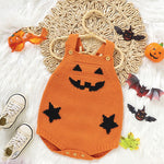 Orange knitted baby romper with jack-o'-lantern face design on a textured surface with Halloween decorations.