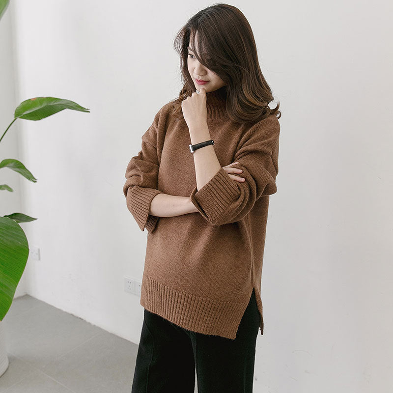 Woman wearing a brown sweater standing indoors with a white wall and plant in the background