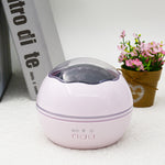 Round purple device with a transparent top on a white surface with a book and flowers in the background